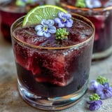 A sophisticated black currant gin cocktail featuring rich berry syrup, fresh lime, and delicate edible flowers.